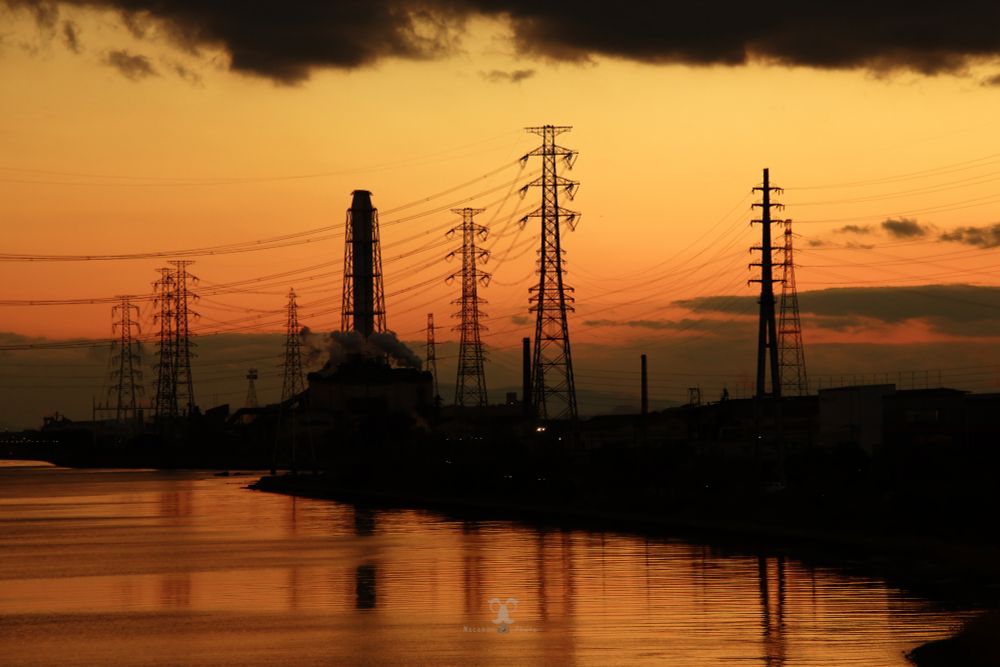 夕焼け空の下、煙を上げる工場と高くそびえる送電線が川沿いに並ぶシルエット風景。水面にはオレンジ色の空が映り込んでいる。