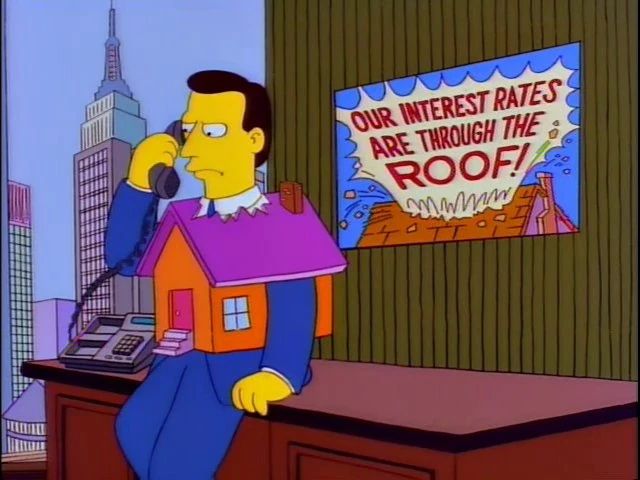 A dark haired man sits on a desk holding a phone. His upper half is encased by a replica house with his head protruding out through the roof of said house. A poster on the wall behind him says “OUR INTEREST RATES ARE THROUGH THE ROOF!”