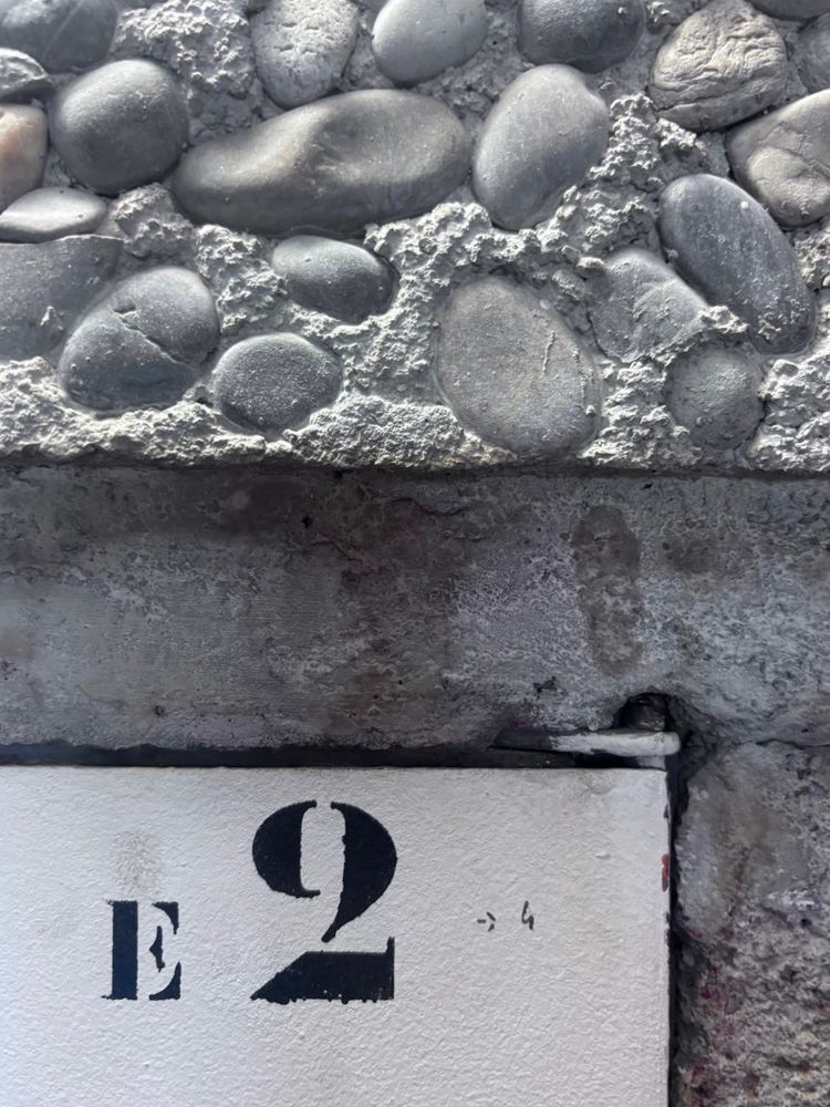 Photo: Close up of a concrete wall. The surface has pebbles and there’s a hint of a door with a stenciled number 2