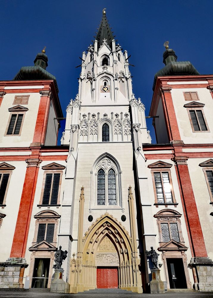 Frontansicht einer Kirche vor dunkelblauem Himmel. Ein Turm aus weißem Stein mit schwarzer Turmspitze in der Mitte, gesäumt von zwei weiteren Türmen mit dunklen Zwiebeldächern, deren Kanten rot ummauert sind. In der Mitte ein großes, spitzbogiges Portal mit roter Eisentür, das von zwei Statuen auf Sockeln gesäumt ist. Links und rechts daneben zwei weitere, kleinere Eingänge. 