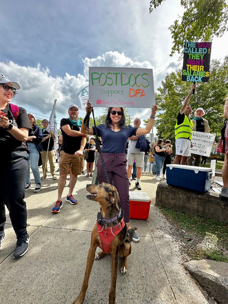 A photo showing a woman holding up a sign that reads “POSTDOCS support DFA #KeepDalStrong”. A happy-looking dog is sitting in front of her and looking back at her. They are at the lockout rally and the crowd can be seen around them.