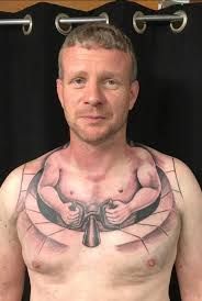 A naked man showing his chest tattoo -ink making it look like he's a tiny body man driving the rest of his body in a little vehicle, like a kuato 