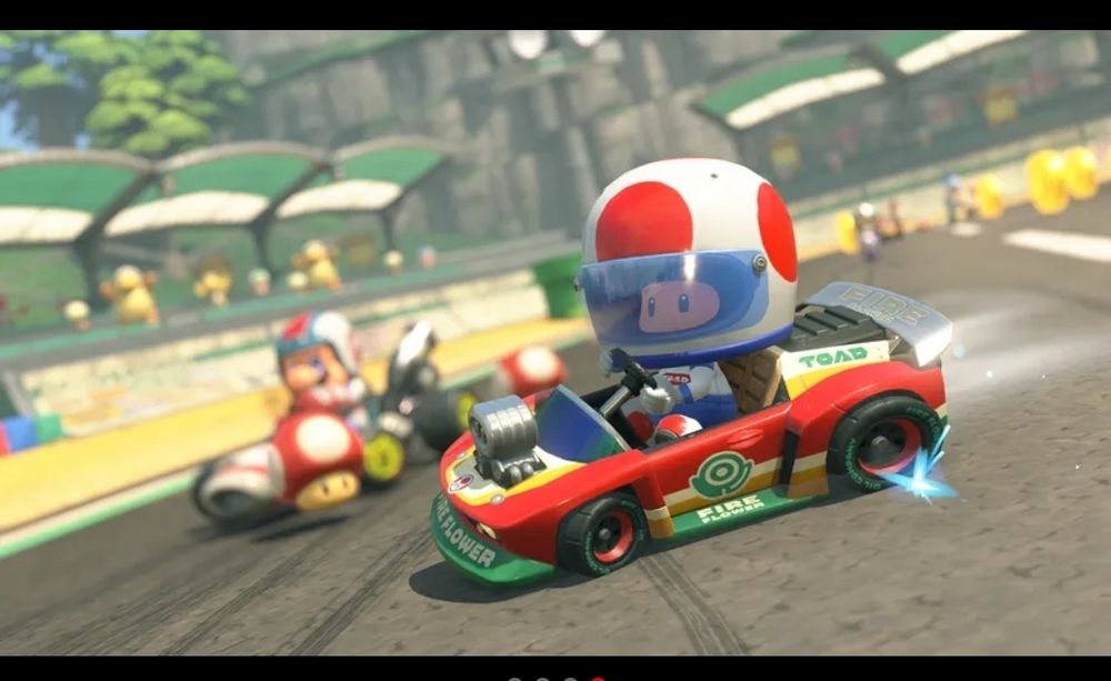 Screenshot from Nintendo Switch 2's Mario Kart World. Toad is racing a red, green, and yellow racecar on a track. He is in a racecar driver outfit. His helmet is his usual white with red circles, but has a blue visor across his eyes. 