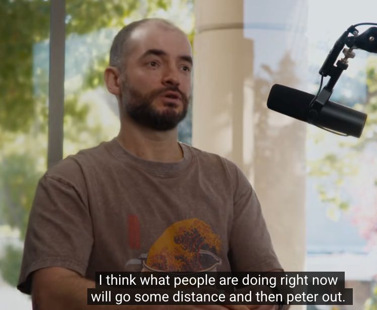 In the image, a man is seated at a podcast table, speaking into a black RØDE microphone on a boom arm positioned to his right. He has a shaved head, a short beard, and is wearing a worn brown T-shirt with an orange-and-yellow graphic. His hands rest loosely in front of him as he talks, and his expression looks earnest and analytical.

Behind him are large windows with bright daylight and green foliage, giving the room a calm, natural ambiance. A tall glass bottle of sparkling water sits on the table near him.

The subtitles at the bottom read:
**“I think what people are doing right now will go some distance and then peter out.”**

