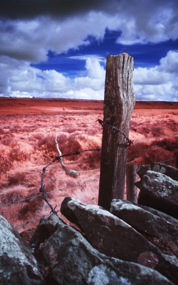 A weathered fence post leaning against an old stone wall. Rusted barbed wire is curled around the post. We can see moorland in the background, with patchy cloud and deep blue sky. This is an infrared photo.