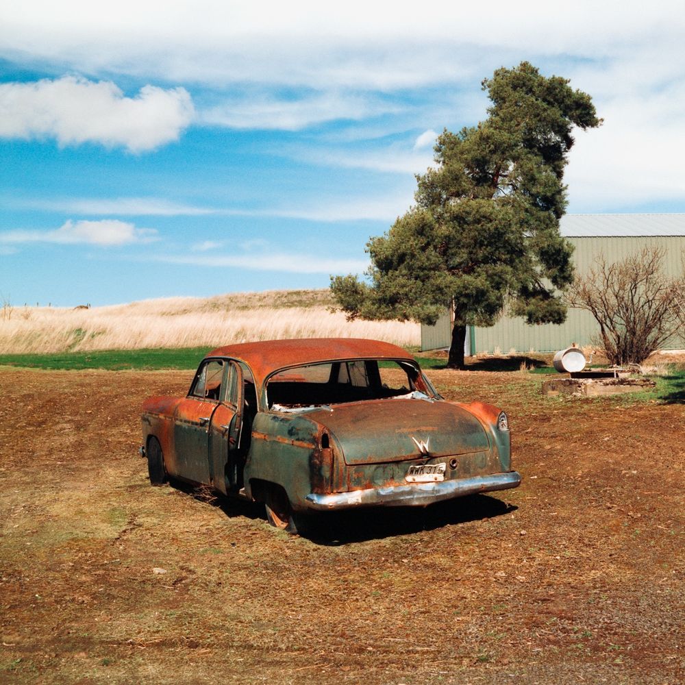 A picture of an old rusty car in a dirt lot with a tree and partially cloudy sky in the background 