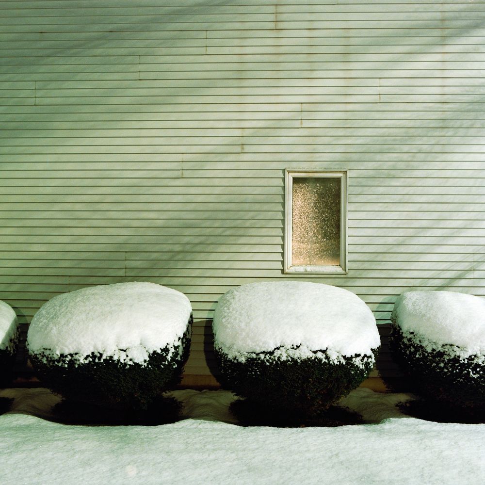 An outer wall with shadow pattern on it and a single window with hedges covered in snow beneath it