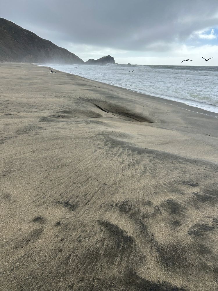 Black and tan sandy beach and wind whipped surf against the rocks. Dark clouds above.