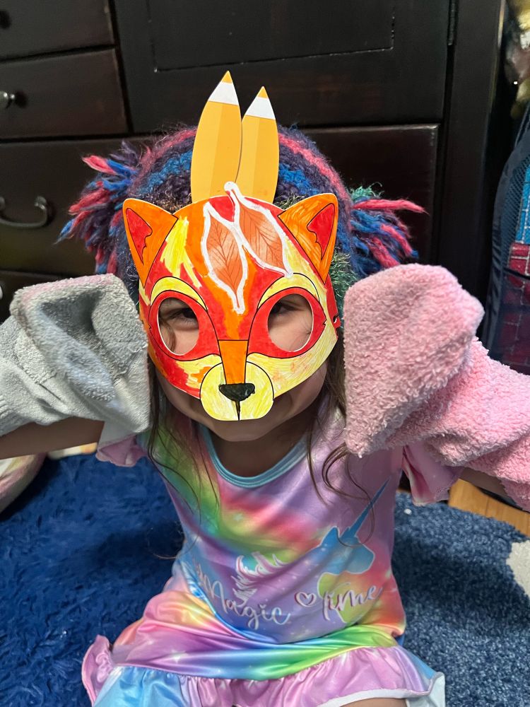 A small child wearing a hand drawn fox mask, magic unicorn pajamas, frilly knit hat, and socks on her hands.