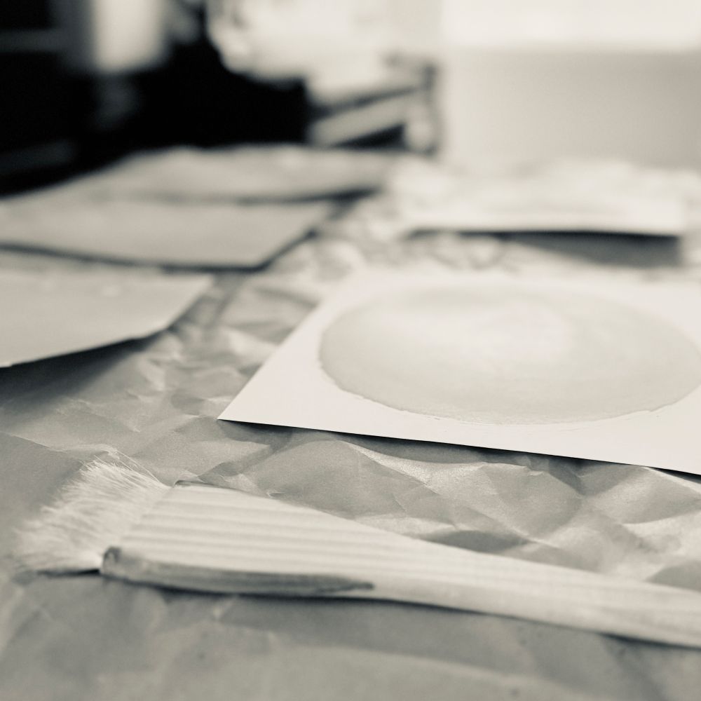 Black and white photo of white rag papers brushed with a light-colored solution, laid to dry on crumpled brown paper, with a broad paintbrush in the foreground. 