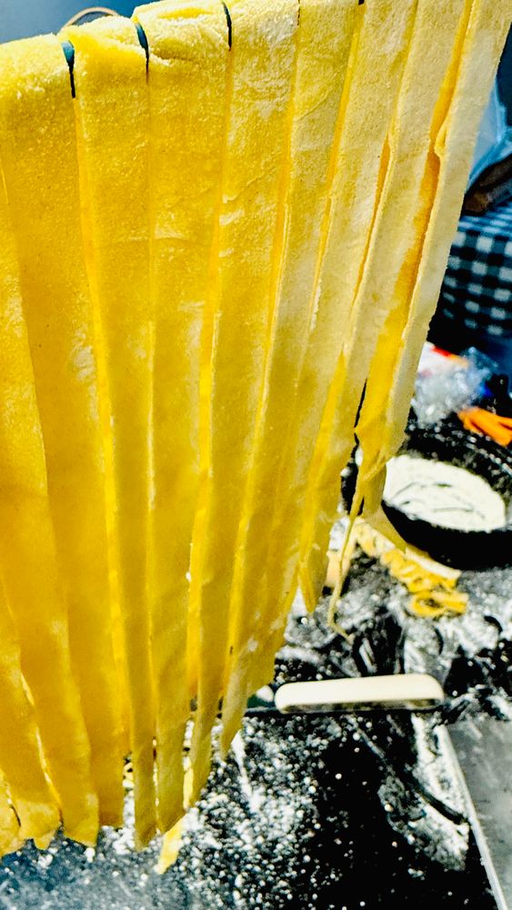 Raw fettuccine-esque pasta hanging down from a wooden rod. An absolute mess of flour and stuff can be seen in the background. 