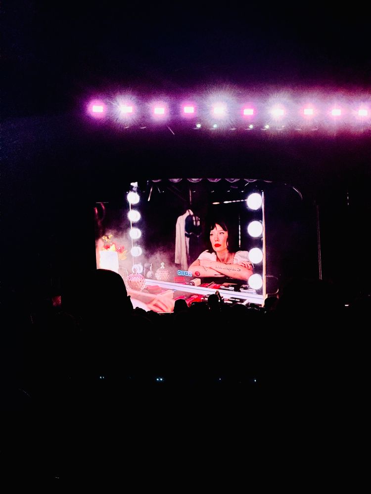The big screen at Halsey’s The Great Impersonator Tour showing them seemingly backstage during the middle of the show. They’re sitting at a lit vanity and staring at themselves in the mirror.