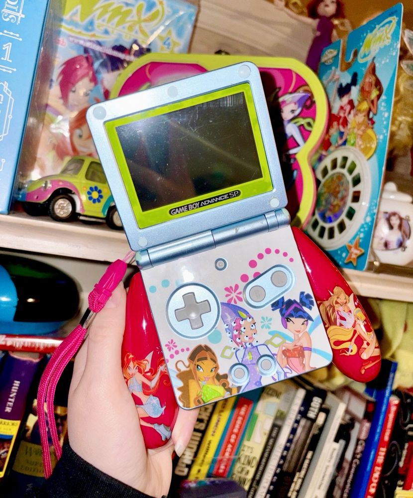 picture of person holding a gameboy advance with winx club decals and a winx club ergonomic attachment set behind a background of a bookshelf with winx club collectibles and books on it 