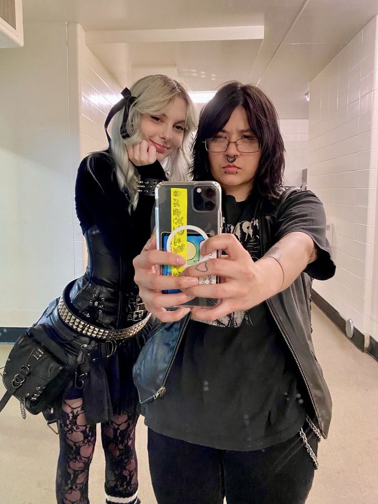 two people posing for a photo in the mirror. person on the left (me) has blue curled hair, is smiling, and is dressed in a black  long sleeve shirt, black corset with buckle details, a black tutu skirt with a studded belt and black purse attached to the hip. accessories include a black bow, black lace tights, spiked bracelets, black and white striped leg warmers. person on the right (viggo) has cool shaggy black hair , a stretched septum and snake bites, cute glasses, and is holding their phone out to take a picture. they are wearing a black cramps black shirt, black vest, and black jeans with a chain. 