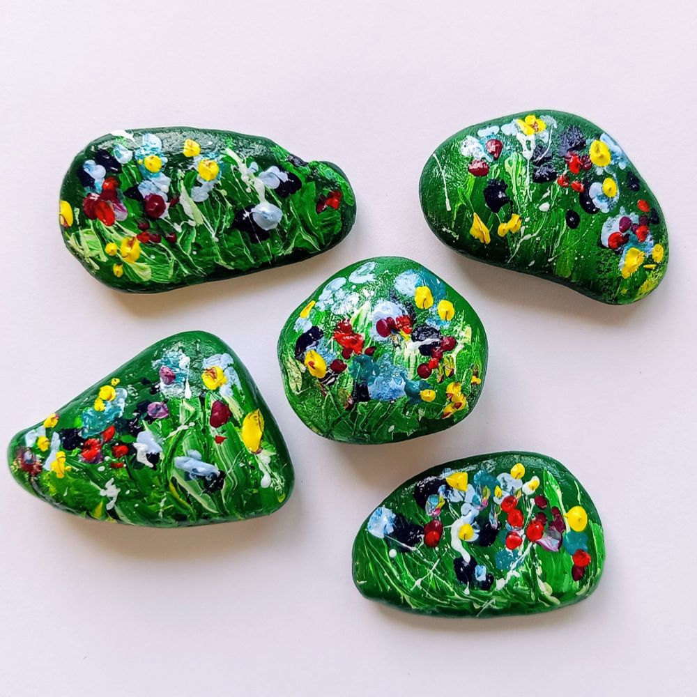 Handbemalte Steine in kräftigem Grün, verziert mit bunten Blumen in leuchtendem Gelb, tiefem Rot, klarem Blau und zartem Weiß. Die Farben wirken lebendig und erinnern an eine blühende Frühlingswiese.