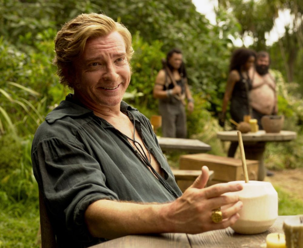 Stede smiling, sitting at a table, his hand stroking a coconut with a straw in it