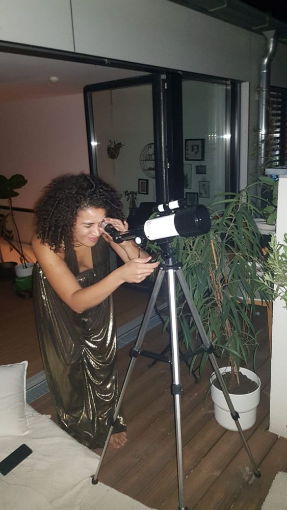 cat in golden party jumpsuit standing on their deck at night, looking through a telescope