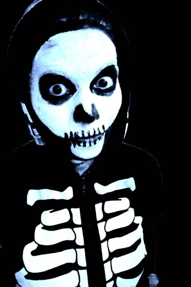 cat dressed up for halloween in a black skelleton onesie, wearing heavy skelleton halloween makeup