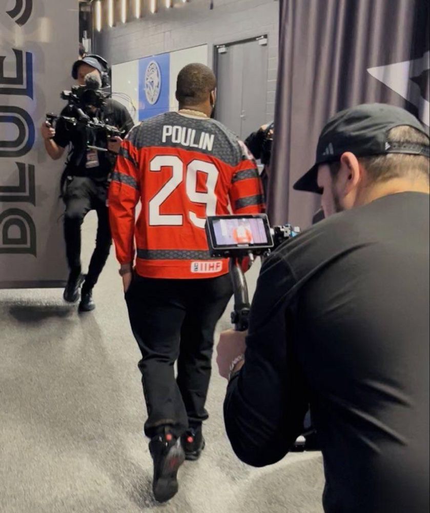 An Afro-Latino man walks away from the camera wearing a red long sleeved hockey jersey with black accents that reads POULIN 29 on the back in white lettering