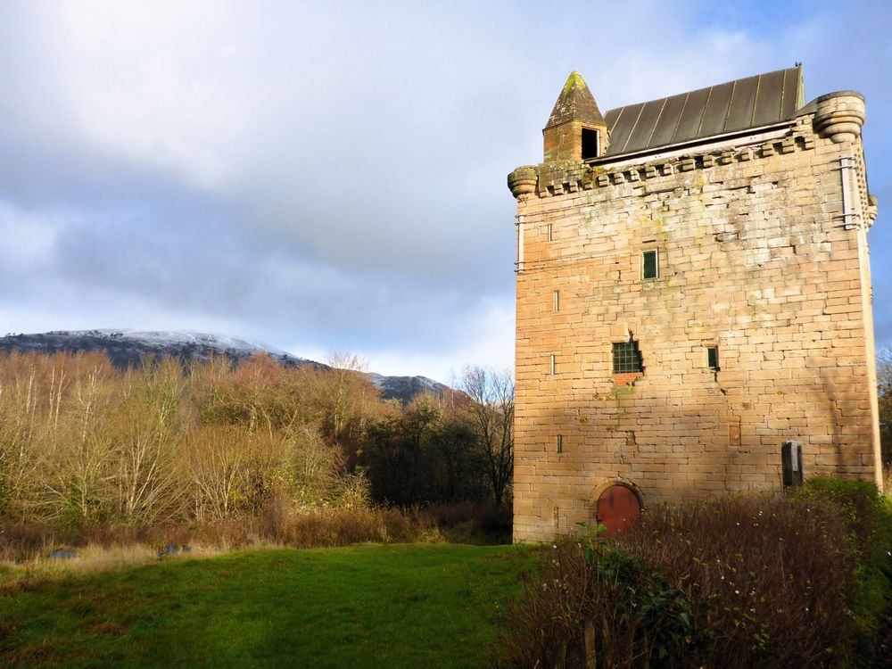 Sauchie Tower, an old, sturdy, rectangular tower, in the light of the afternoon sun. On the left bare shrubs and trees, in the distance hills with a dusting of snow on the tops.