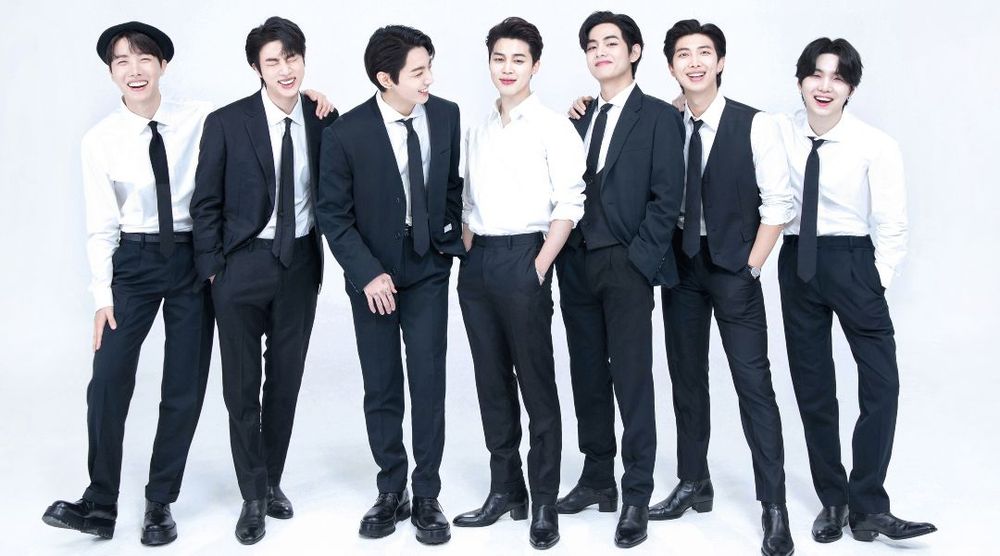 BTS members wearing various combinations of suits, shirts, ties, and slacks. From L to R: Hobi, Jin, Jungkook, Jimin, Taehyung, Namjoon, and Yoongi. They are all smiling or laughing and look so happy. 