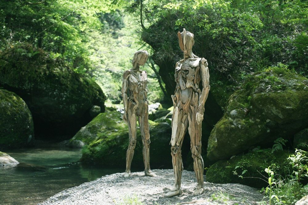 The sculptures and images are by Nagato Iwasaki (IG: nagato_iwasaki).

Within a green forest, thick with trees and boulders, stands two wooden sculptures crafted from asymmetrical pieces of wood. They stand upon a massive rock along the shore of a clear stream weaving through the woods and rocks.