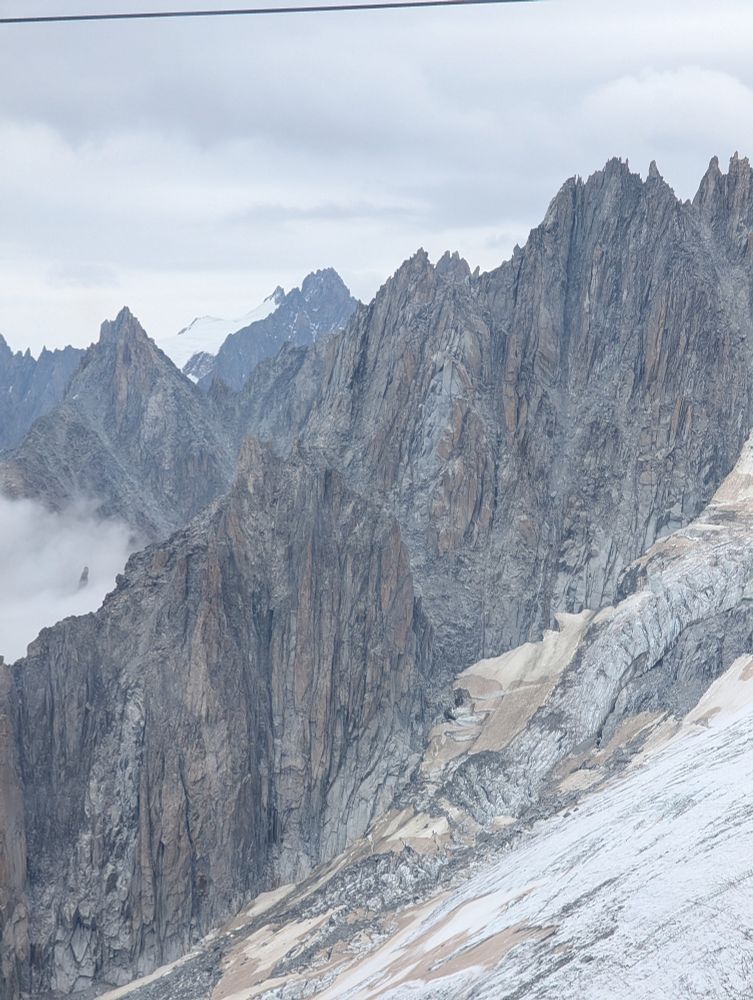 Picture of a mountain range in the French Alps that has many vertical striations. There are two layers of mountains, clouds in the left middle. The lower right is a glacier field. 