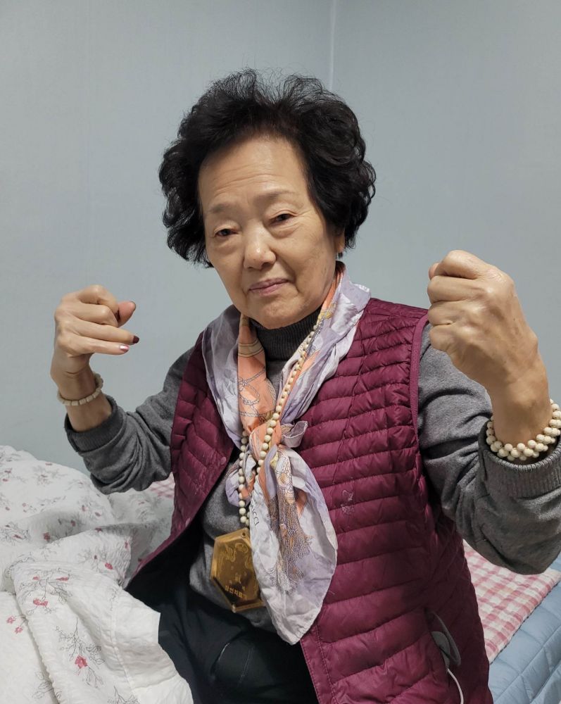 Hyun Sook's mom flexing her muscles while wearing a "health medallion" made of plastic and gold paint that she bought from a conman for 4000 dollars.