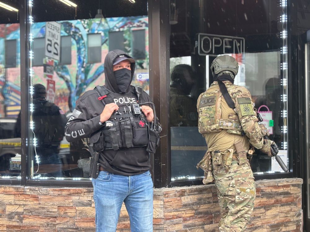 This time a man in a gray hoodie, baseball cap and mask to hide his identity. This guy has POLICE emblazoned across his chest armor. He and his camo-wearing asshole friend are both armed with guns.