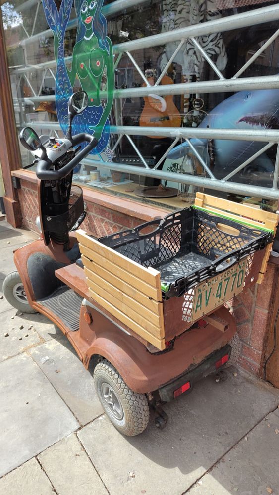A rusty looking scooter with a timber / crate rear basket, NC plate, in front of a store with aluminium bars across the window. There are bricks under the window in a decorative bond, and there is a small key safe at one end of the brickwork.
The window is decorated with a mermaid in a hippy style.