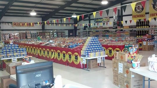 Interior of a fireworks retailer with a substantial array of fireworks on sale. Banners on the walls read “FIREWORKS”. The main stand is branded Black Cat.