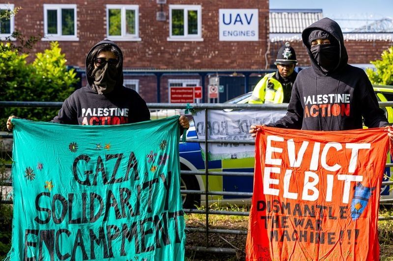 Two people wearing black hoodies and masks stand outside the gates to a factory. One of the two is wearing sunglasses. The visible part of the factory is a brick 2 storey building with white PVC windows. The two people are holding hand made banners and the words on their hoodies read "PALESTINE ACTION. Inside the factory gates are a police vehicle and a uniformed police officer. 

There is another banner attached to the steel gate.

The visible banners read "GAZA SOLIDARITY ENCAMPMENT" and "EVICT ELBIT / DISMANTLE THE WAR MACHINE". The sign on the factory is blue text on white and reads "UAV ENGINES".

No genocide is depicted.