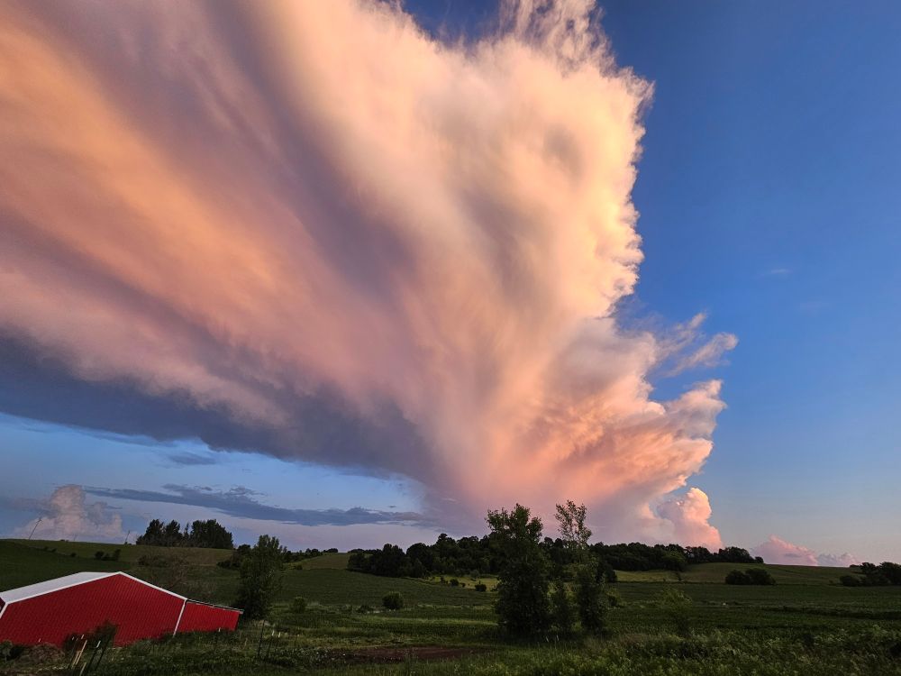 Landscape with red barn and fields is overshadowed by large columns of clouds in ivory, pink, and grey colours caused by the sunset