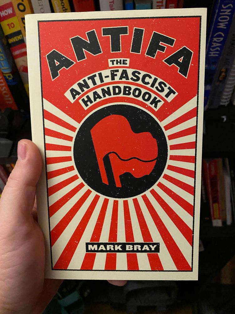 A photo of a hand holding a paperback copy of Mark Bray’s “Antifa”. Behind it is a bookshelf full of other books.