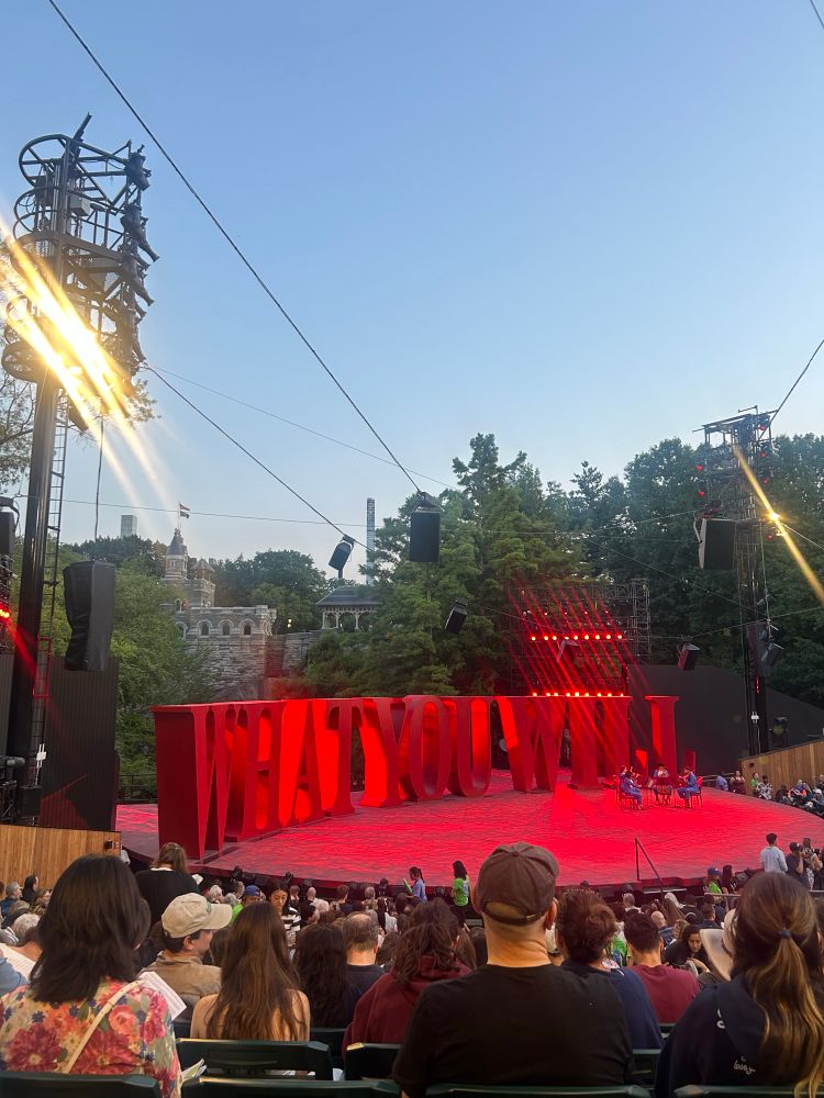 the delacourte theater stage at sunset, on stage in red light read large words that say “what you will”. There is a string quartet on stage and people sitting in the rows ahead of me. The sky is light blue and Central Park is scene in the background of a stage. 