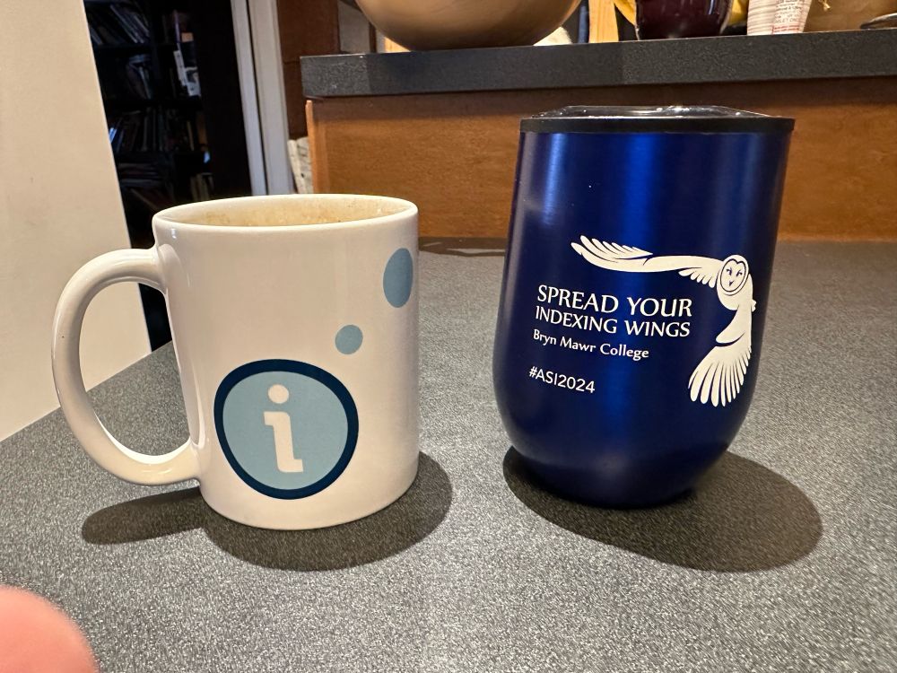 Two mugs. White one showing logo for Indexing Society of Canada. Dark blue tumbler from ASI’s 2024 conference. 
