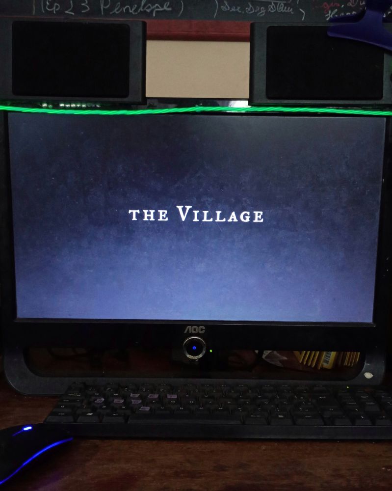 Foto do meu computador exibindo o letreiro do filme The Village no centro da tela, todo em maiúsculas, em fonte clássica e em branco, estilizado com a letra V mais alta do que as demais. Fundo preto. Há caixas de som retangulares acima da tela e um cabo iluminado verde esticado também acima. Parte de um mouse com iluminação colorida e o teclado estão à frente sobre mesa de madeira escura. Está um pouco claro e na parede ao fundo há parte de um quadro de giz com coisas escritas.