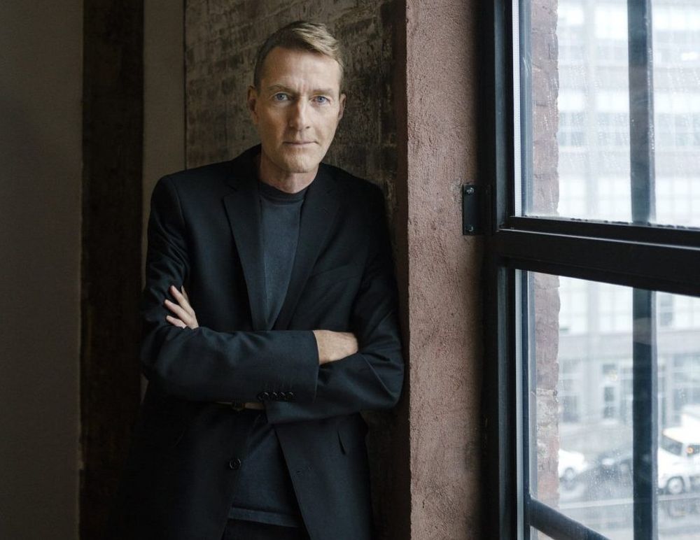 A photo of author Lee Child standing next to a window with his arms folded and a wry expression. He does not look a day over sixty