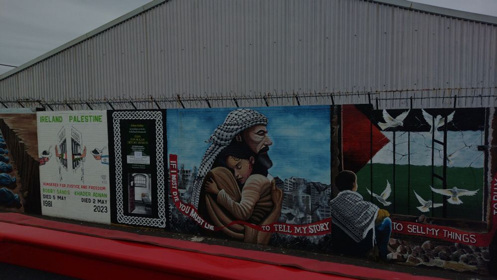 Central mural is a man wearing a keffiyeh, hugging a child, with a destroyed building in the background. 