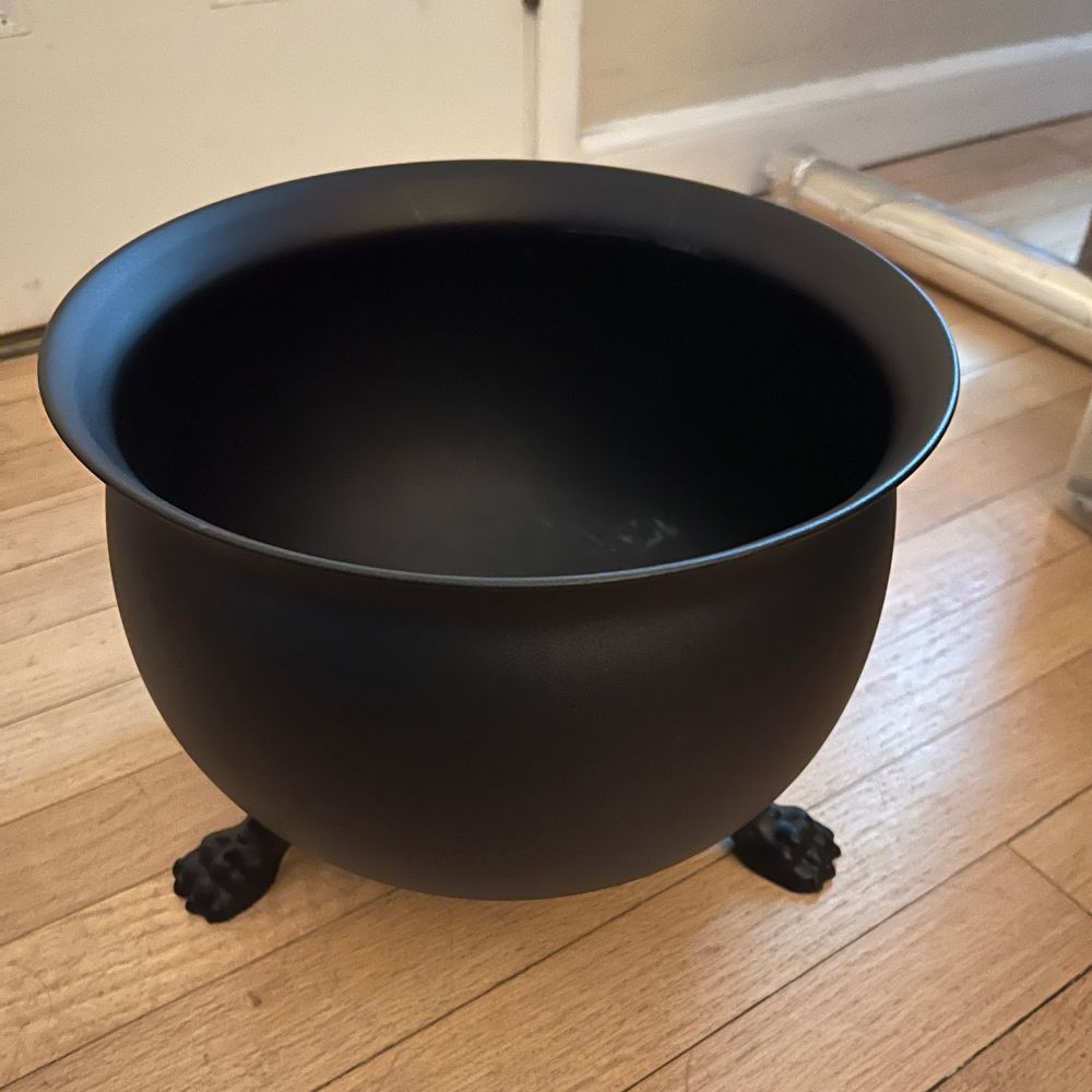 Black metal cauldron with feet (paws, really) on a wooden floor. It is about a foot tall and about 16 inches in diameter. 