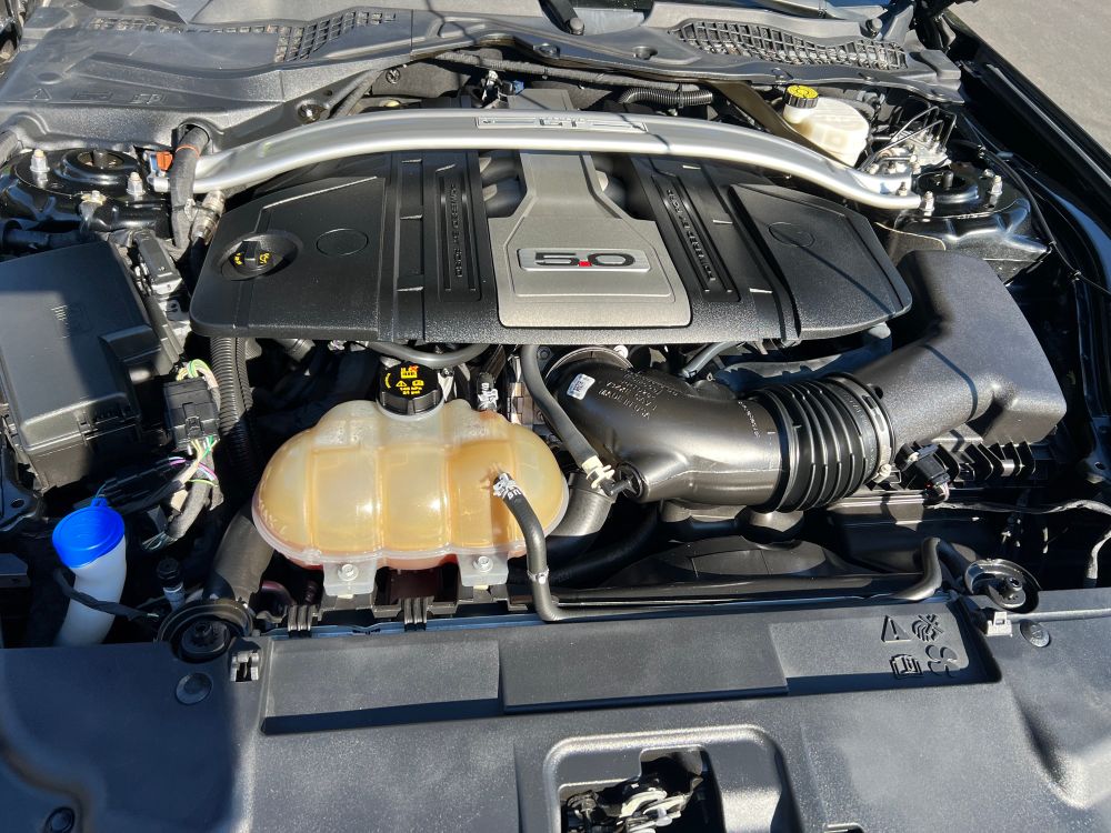 A very clean Coyote 5.0L engine that makes 460hp on 91 octane gas. The engine has a cover, and the coolant reservoir and cold air intake are prominent. 