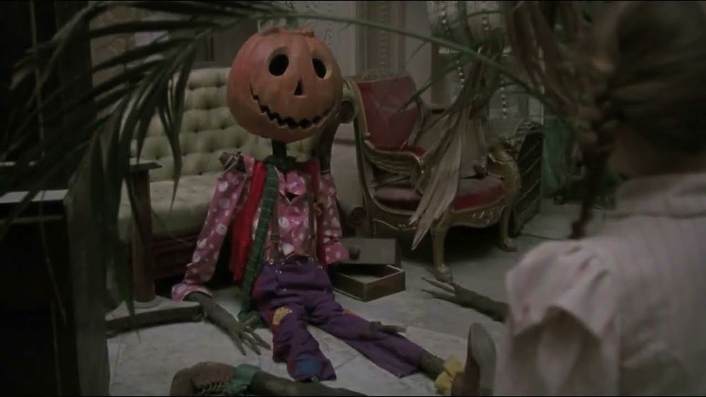 Jack Pumpkinhead from the movie Return To Oz. He is a scarecrow with spindly stick legs, a jack o’lantern head carved with a smile, and mismatched patterned clothes. He sits among various household junk and discarded furniture. The back of Dorothy’s head is visible on the right side of the frame.