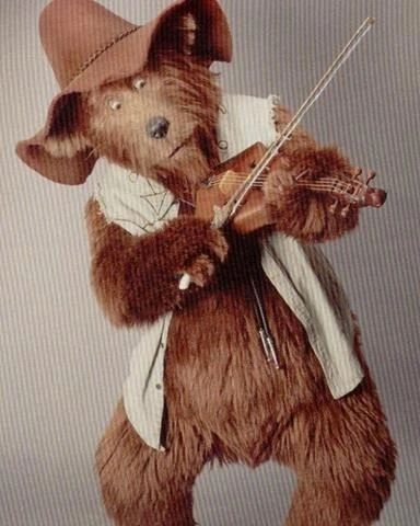 Zeb Zoober from The Country Bears (2002). He is a brown bear in a beige vest, floppy brown hat, and bolo tie, and he is playing the fiddle.