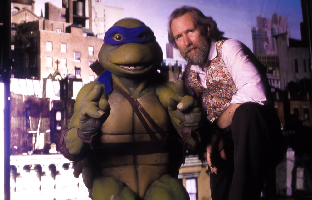 Jim Henson in a floral vest posing with Leonardo the Teenage Mutant Ninja Turtle from the 1990 movie against a cityscape backdrop.