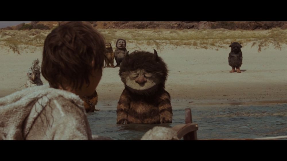 A still from Where The Wild Things Are (2009). Max sits in a boat with his back to the camera. Carol, a large monster with a brown striped torso and small horns, stands and looks dejected in the water. Other various monsters look on from the shore.