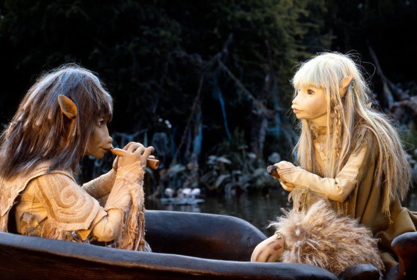 A still from The Dark Crystal. Jen, Kira, and Fizzgig sit in a boat sailing through a marsh. Jen is playing his flute while Kira is rowing the boat.