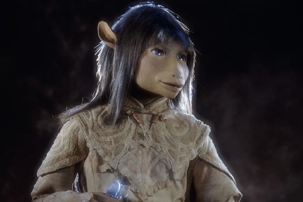 Jen from The Dark Crystal. He is a humanoid elf creature with tan skin, blue eyes, and dark hair that has blue streaks in it. He wears layered beige clothes and holds the crystal shard in his hand. He is looking wistfully off camera with a slight smile.