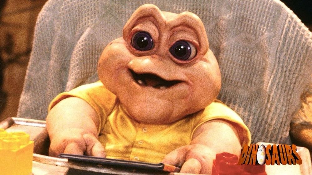 Baby Sinclair from the TV show Dinosaurs. He is a small pink dinosaur in a yellow onesie with large brown eyes. He sits in a high chair against a light blue blanket.