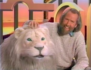 A still from The Jim Henson Hour. Jim Henson, in a grey sweater and smiling, sits with a white lion with green eyes against a geometric CGI background. 