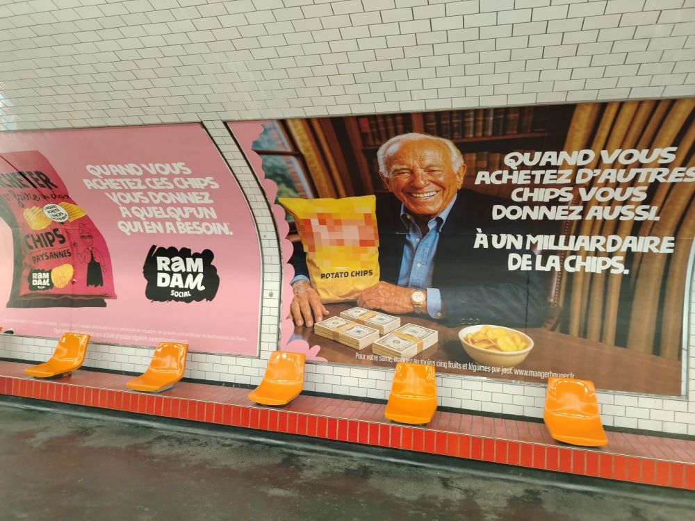 2 panneaux publicitaires dans le métro parisien : sur le 1er un paquet de chips de la marque Ramdam Social "quand vous achetez ce paquet de chips, vous donnez à quelqu'un qui en a besoin". Sur le 2e, un business man au cheveux blancs devant un paquet de chips de marque masquée "quand vous achetez d'autres chips, vous donnez aussi. À un milliardaire de la chips". 